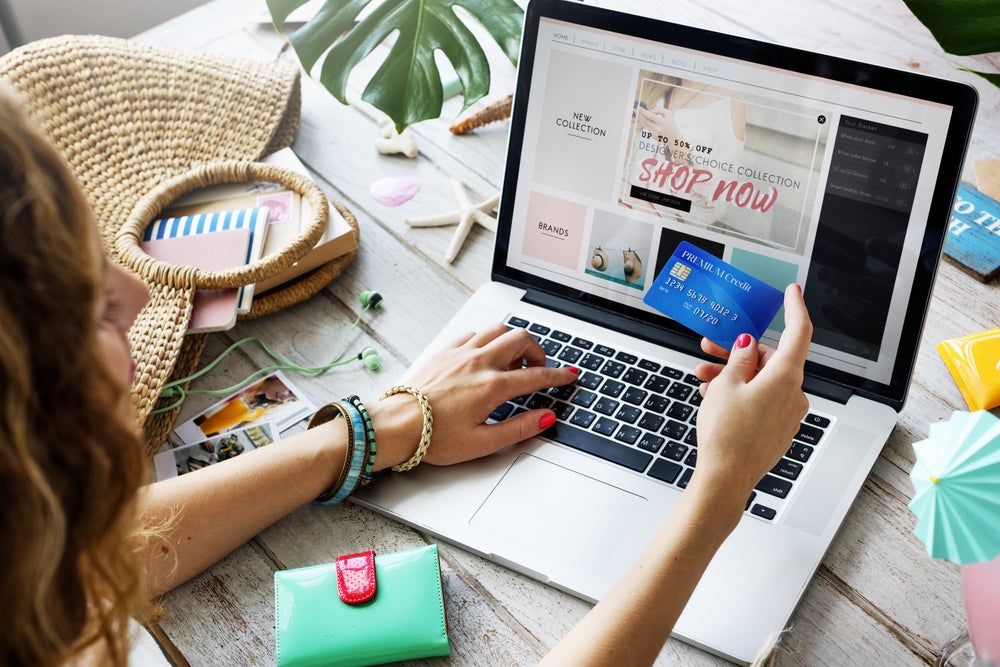 Person shopping online on a laptop, holding a blue credit card, with beach-themed accessories nearby.