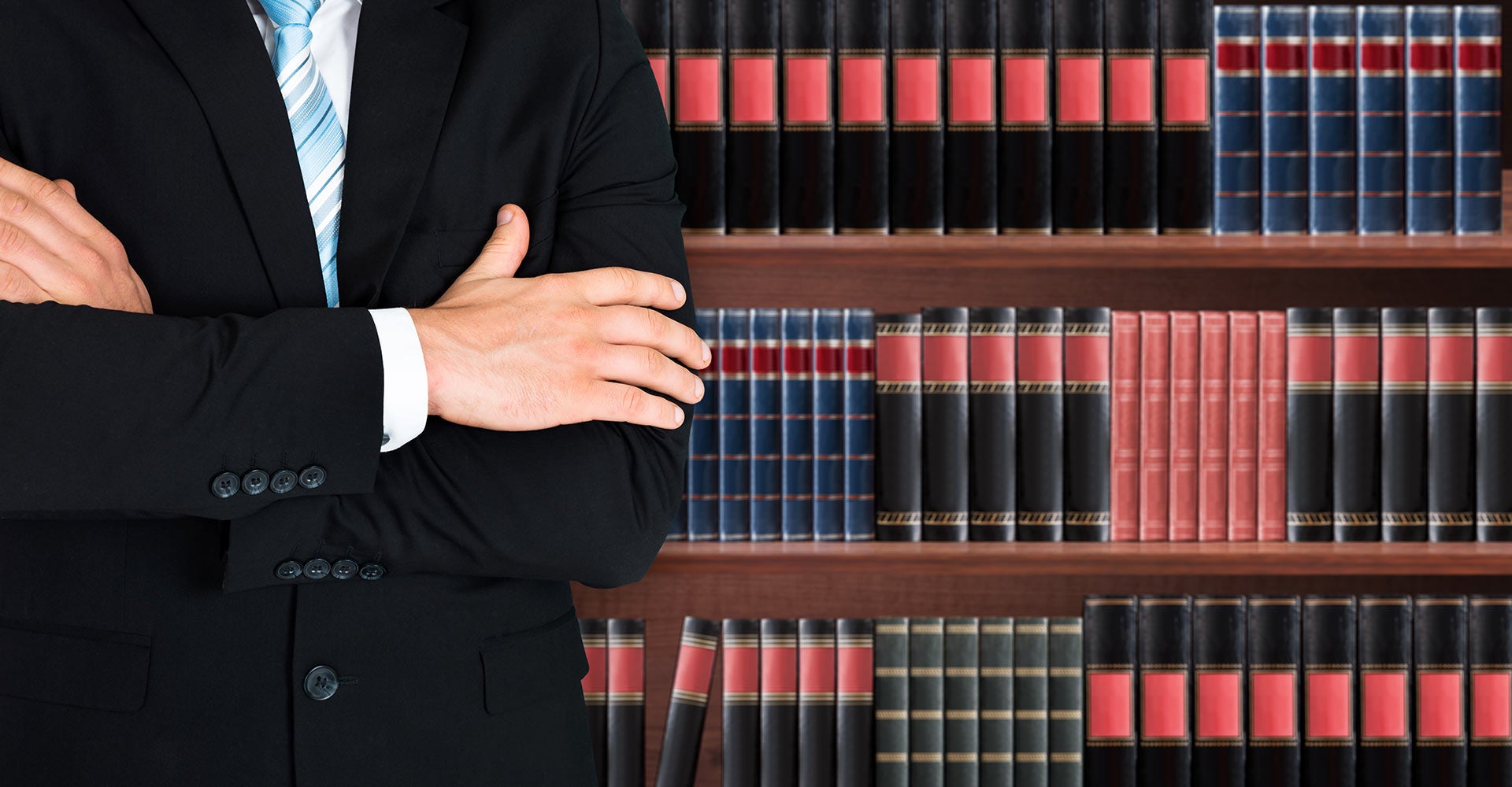 Torso of a man in a black suit and blue tie with arms crossed, in front of a wooden bookshelf filled with books.