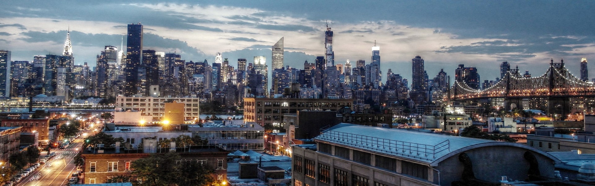 Skyline of a metropolis with brightly lit skyscrapers and bridges under a dramatic sky at twilight.