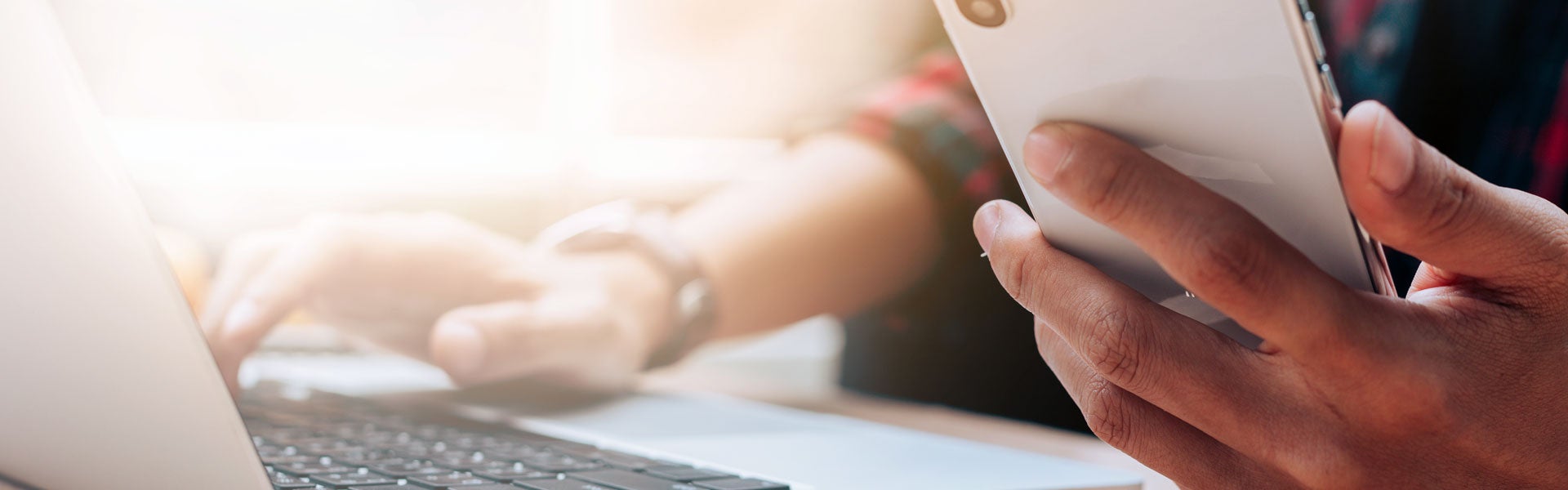 Close-up of a person holding a white smartphone in one hand while the other types on a laptop keyboard.