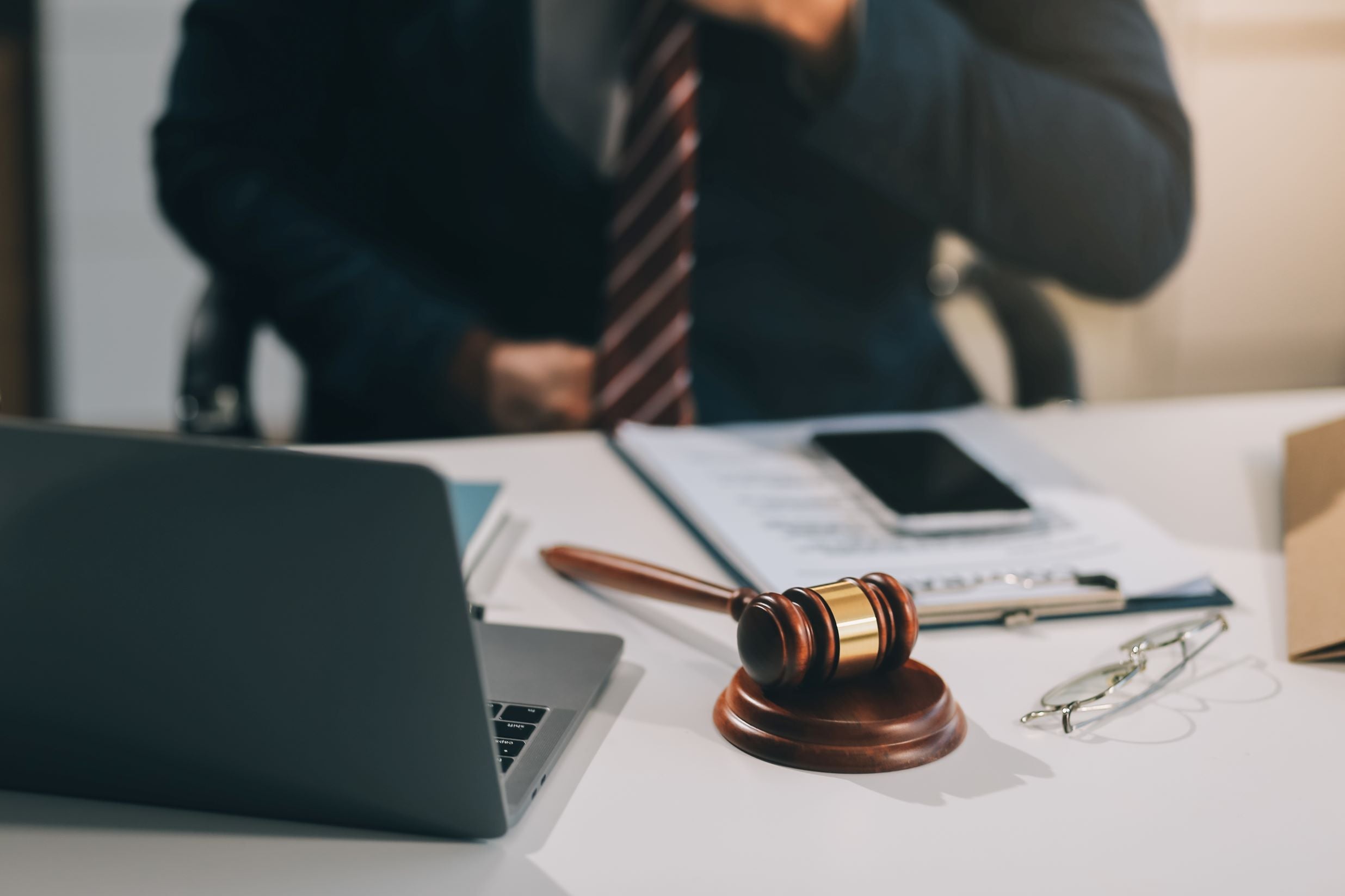 Judge gavel and legal documents on a white desk with a laptop and a man in a suit in the background.