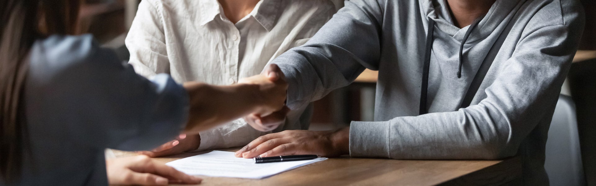 Three people shaking hands across a table with a document and pen on it, sealing a deal.