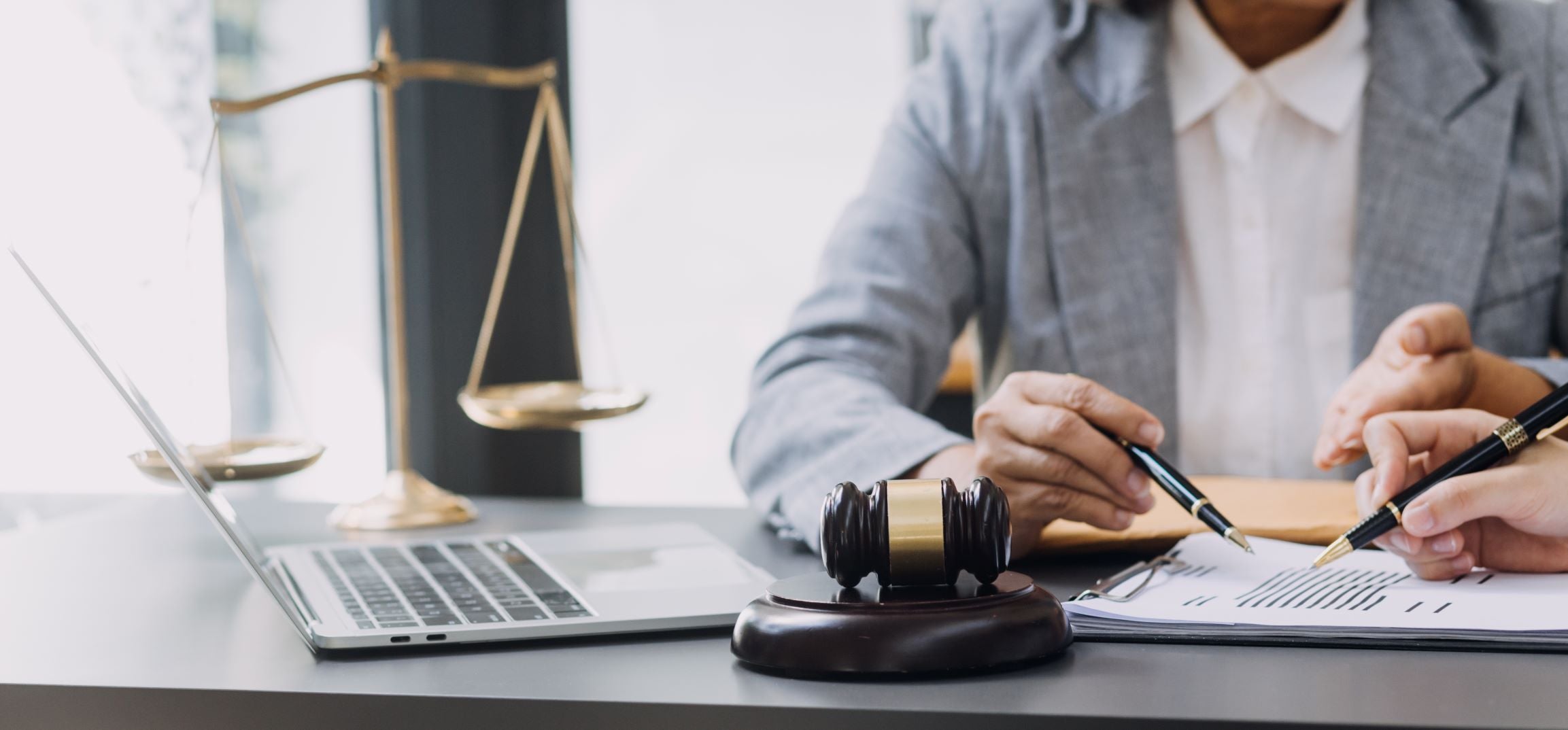Two people review documents at a desk with a laptop, gavel, and scales of justice.
