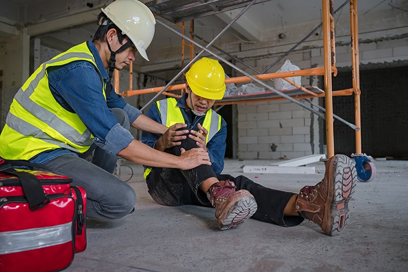 A construction worker in a yellow hard hat clutches his knee, in pain on the ground, assisted by another worker.