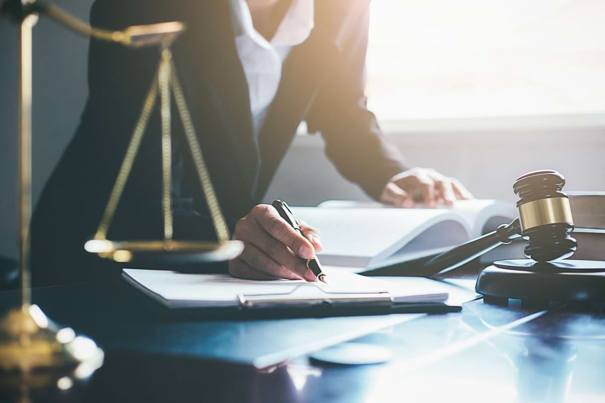 A person in a suit writes on paper next to large books, a gavel, and a golden scale of justice.