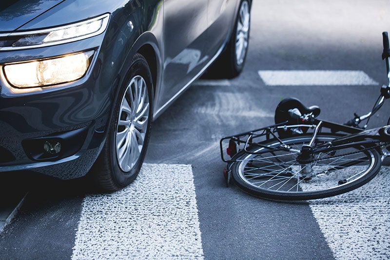 A dark gray car with headlights on, parked next to a fallen bicycle on a crosswalk, indicating an accident.