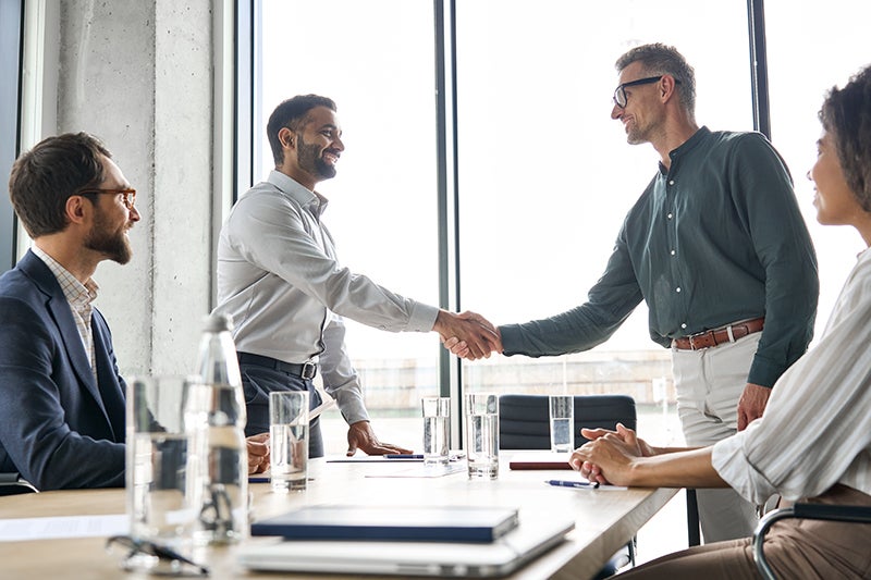 Two men shaking hands across a conference table with other colleagues present, signifying a business agreement.