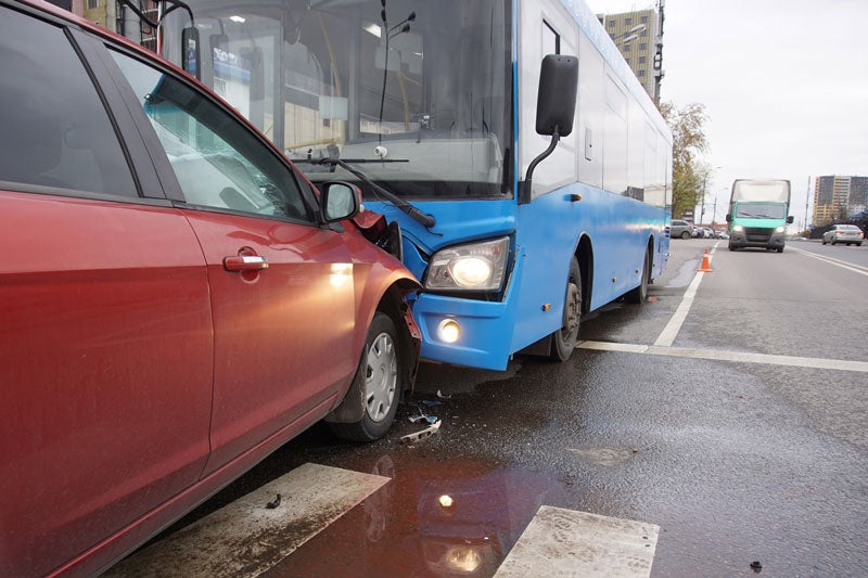 A red car and a blue bus demonstrate the aftermath of a collision on a wet city street.