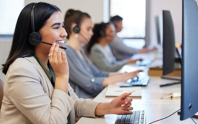 A smiling call center agent, wearing a headset, types on a computer in a row with other agents.
