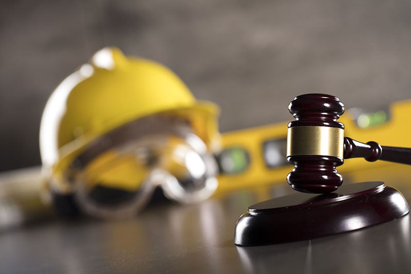A gavel and sound block in the foreground with a yellow hard hat, safety glasses, and level blurred in the background.