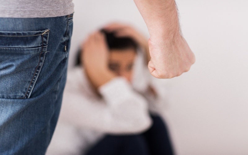 A man's clenched fist and leg in jeans in the foreground, with a blurred, fearful person in the background.