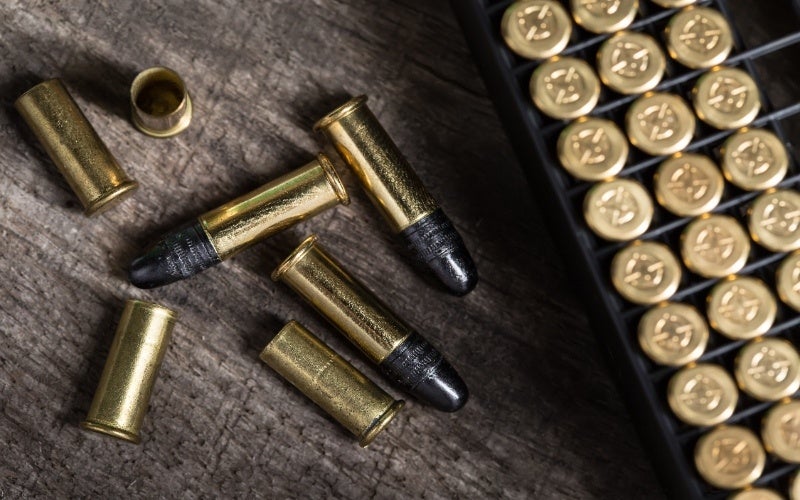 Close-up of golden rifle cartridges with black tips on a wooden surface, next to an ammo box.