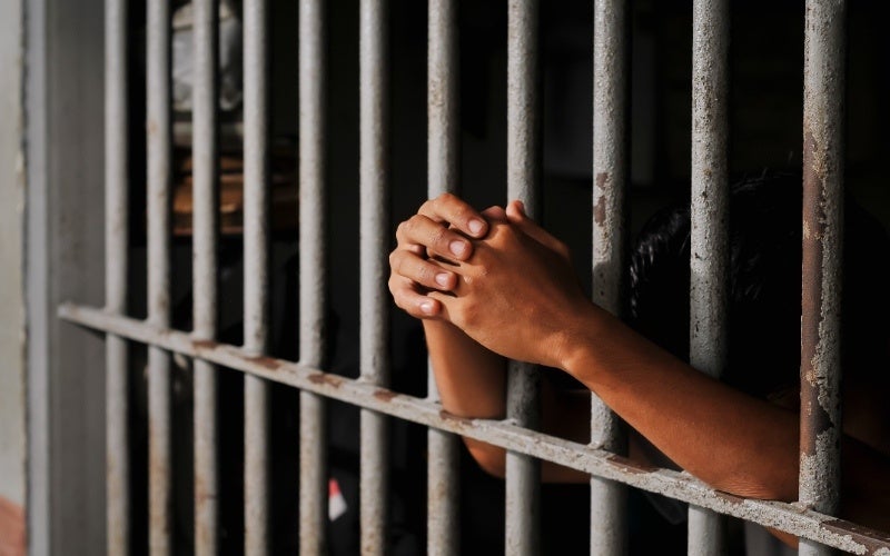 A person's clasped hands are visible through the rusted bars of a jail cell, suggesting imprisonment or detention.