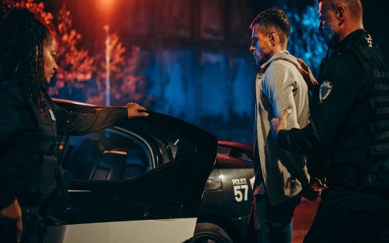 Two police officers apprehend a man in handcuffs next to an open squad car at night.