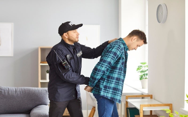 A police officer in uniform handcuffs a man in a plaid shirt inside a home.