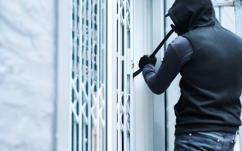 A person wearing a black hoodie and vest uses a crowbar to pry open a door with a security gate.