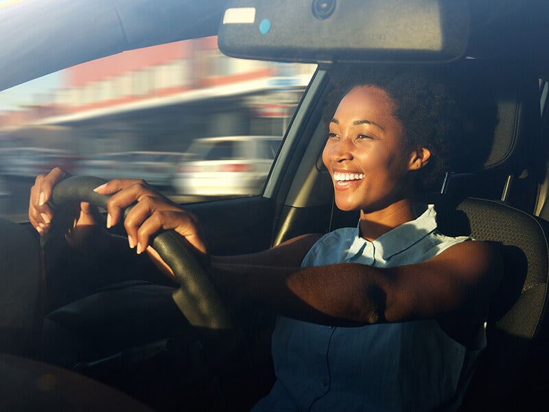 A smiling Black woman driving, hands on the steering wheel, with blurred motion of passing cars and sunlight.
