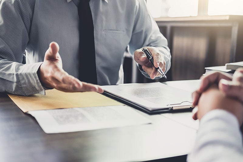A person in a striped shirt and tie discusses documents on a table with another person.