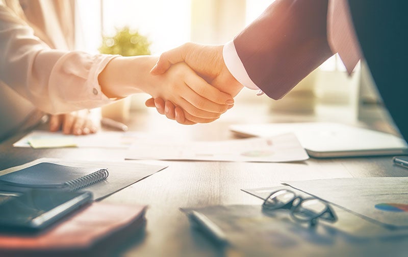 Two people shake hands over a business table with papers, glasses, and a notebook on it.