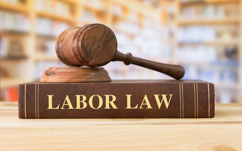 A wooden gavel rests atop a brown book titled LABOR LAW in gold letters, with a blurred bookshelf background.
