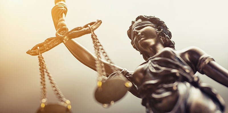 A low-angle shot of a bronze Lady Justice statue with a blindfold, holding balanced scales, bathed in bright sunlight.
