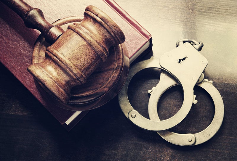 A wooden gavel resting on a red book, with silver handcuffs on a dark table, symbolizing law and justice.