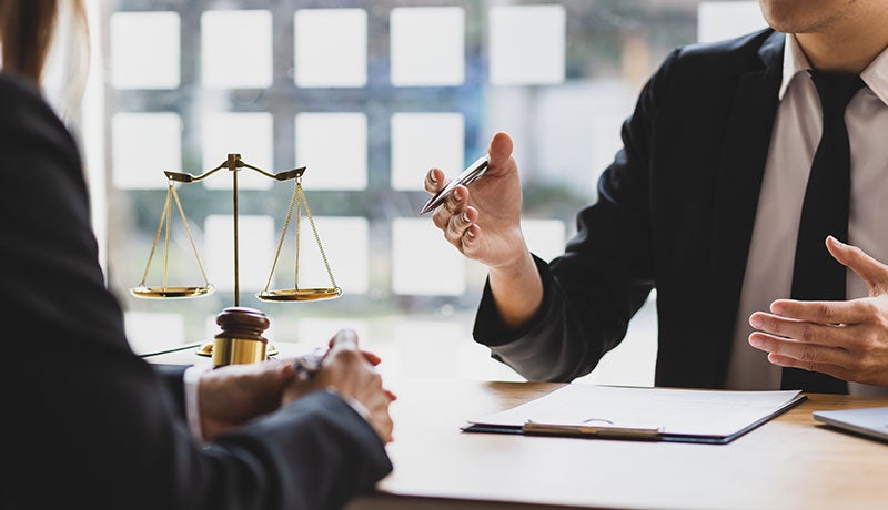 Two people, dressed in suits, discuss legal matters at a table with scales of justice and paperwork.