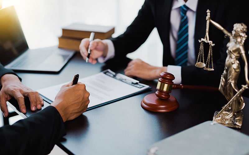 Two people at a desk: one signing a document, another pointing, with a gavel and Lady Justice statue visible.