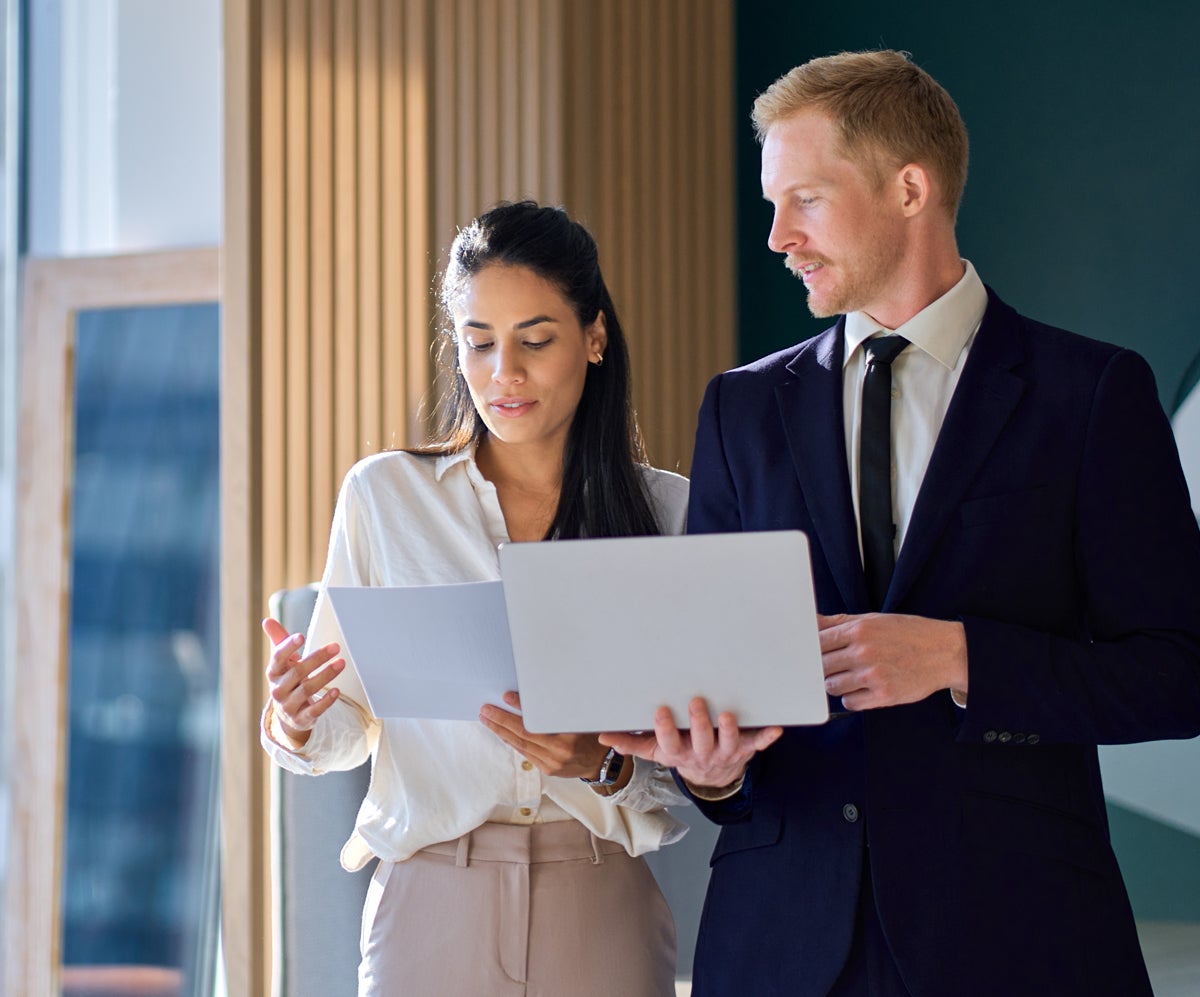 Two professionals collaborating: a woman holding documents and a man holding a silver laptop, both looking at the papers.