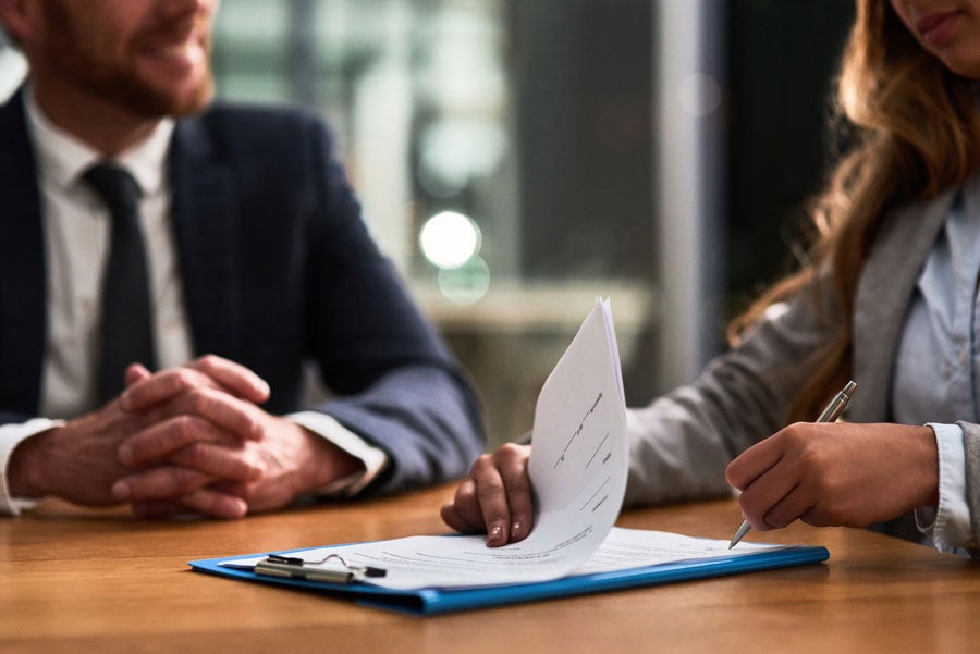 A person in a suit observes as another person's hand signs a document on a blue clipboard.