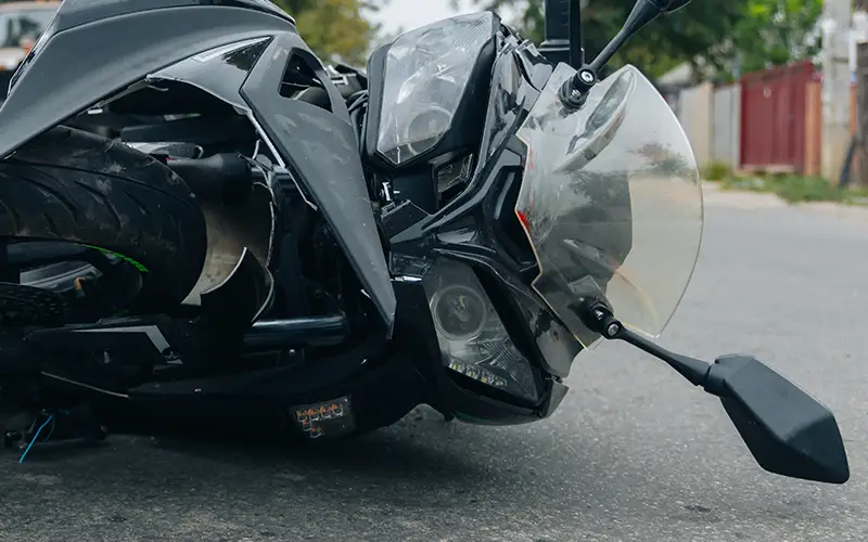 A black motorcycle lays crashed on its side on gray asphalt, front wheel toward the viewer, with bent bodywork.