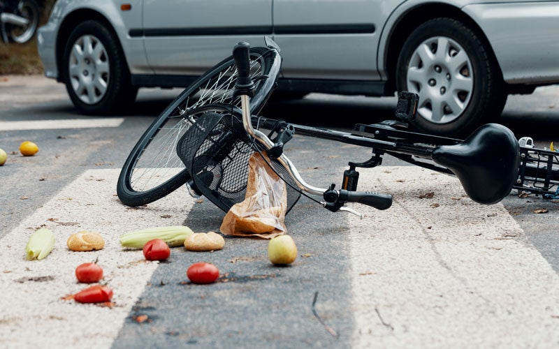 A bicycle and groceries spilled on a crosswalk with a silver car in the background, implying an accident.