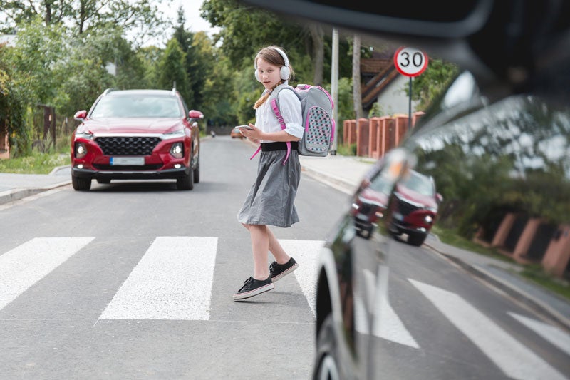 A girl with headphones, a backpack, and a phone crosses a street in a crosswalk as a red car approaches.