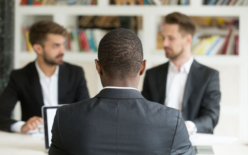 Black man seen from behind in an interview with two white men in suits.