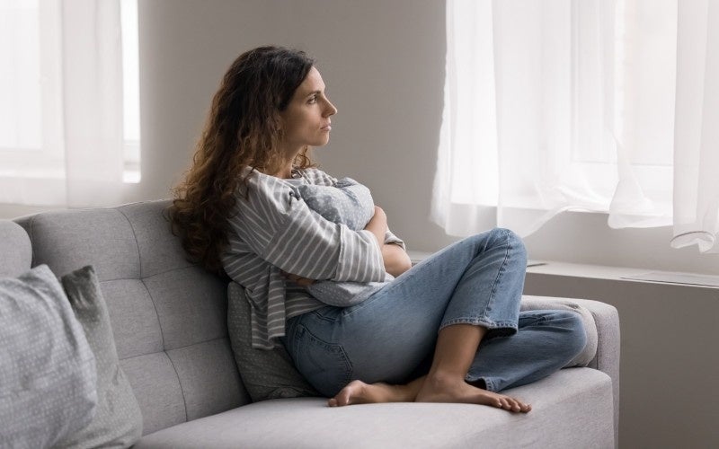 A pensive woman with curly hair sits on a gray sofa, hugging a pillow and looking out a window.