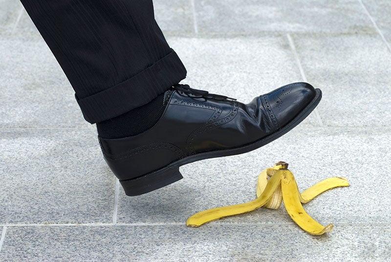 A man's polished black shoe is about to step on a yellow banana peel on a grey tiled sidewalk.