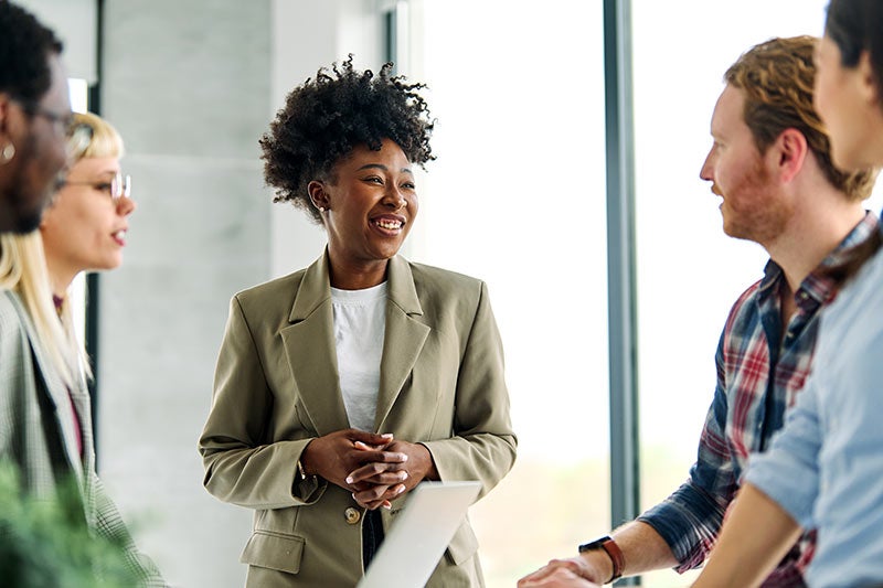 A smiling Black woman in a business suit speaks to diverse colleagues during a meeting in a bright office.