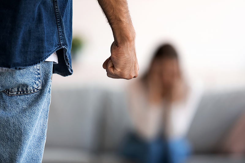 A man's clenched fist in focus in the foreground, with a blurred, distressed woman covering her face in the background.