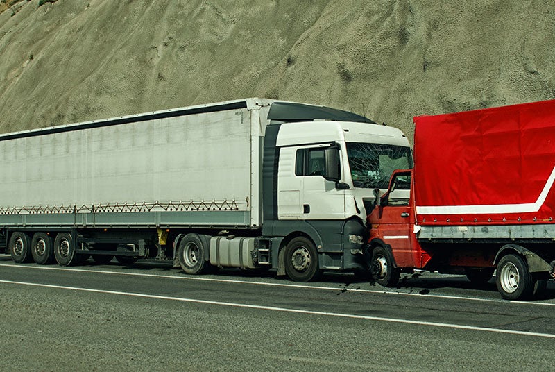 A white semi-truck with a crushed windshield collided head-on with a red truck, on a road by a rocky slope.