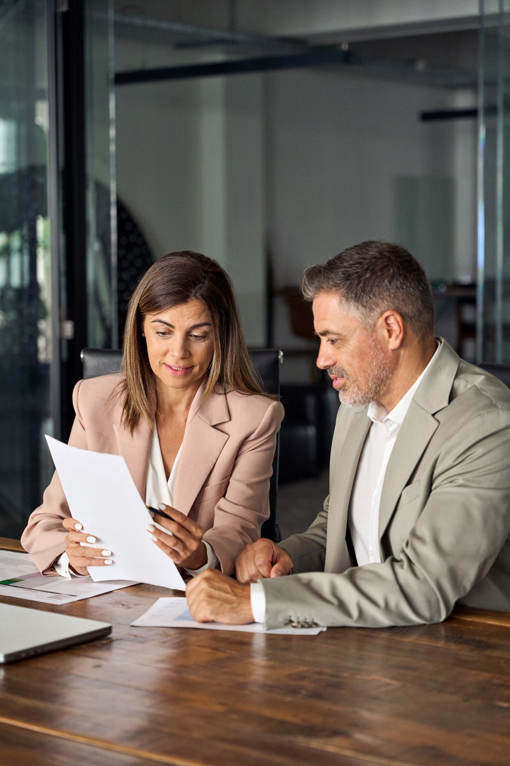 A woman in a rose blazer holds papers while a man next to her in a suit looks on, both seated at a dark wooden table.
