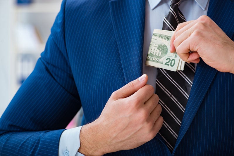 A man in a blue pinstripe suit and striped tie discreetly places a wad of twenty dollar bills into his jacket pocket.