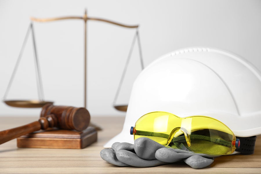 Judge's gavel, scales of justice, hard hat, safety glasses, and work gloves, representing legal aspects of workplace safety.