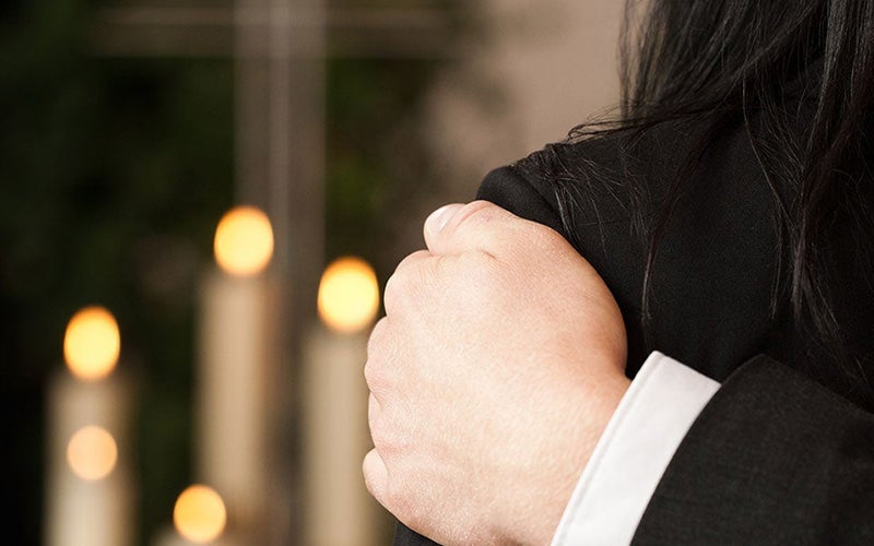 A hand rests gently on a grieving person's shoulder, with blurred flickering candles in the background.