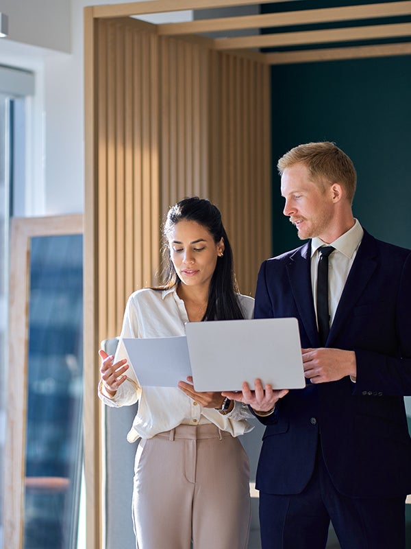 Two business professionals, a woman and a man, standing and looking at a laptop and document together.