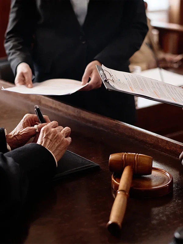 A person signs a document at a wooden table in a courtroom, a gavel lies nearby.